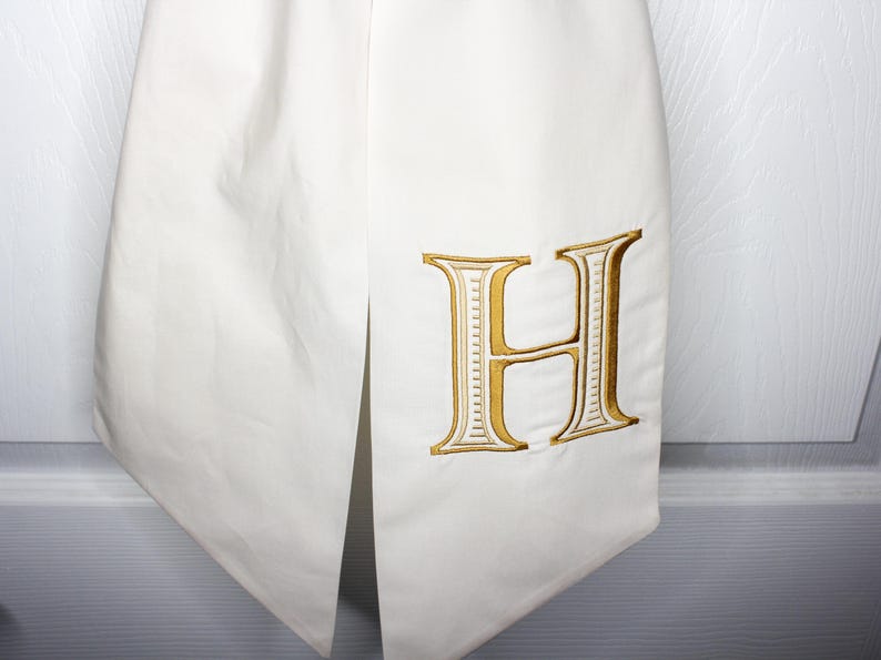 May include: A white fabric item featuring a large, embroidered letter H in a metallic gold thread with white detailing. The fabric has a smooth texture and a vertical slit, showcasing a clean, structured design with precise stitching on the letter.