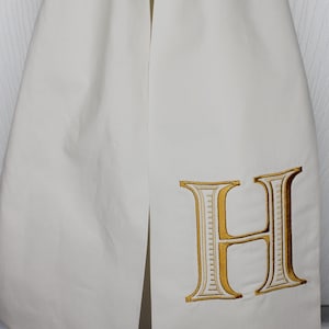 May include: A white fabric stole featuring a large, embroidered letter H. The letter is stitched in a metallic gold thread with a white interior fill and a dark outline, creating a bold, decorative monogram detail on the smooth, solid white textile.