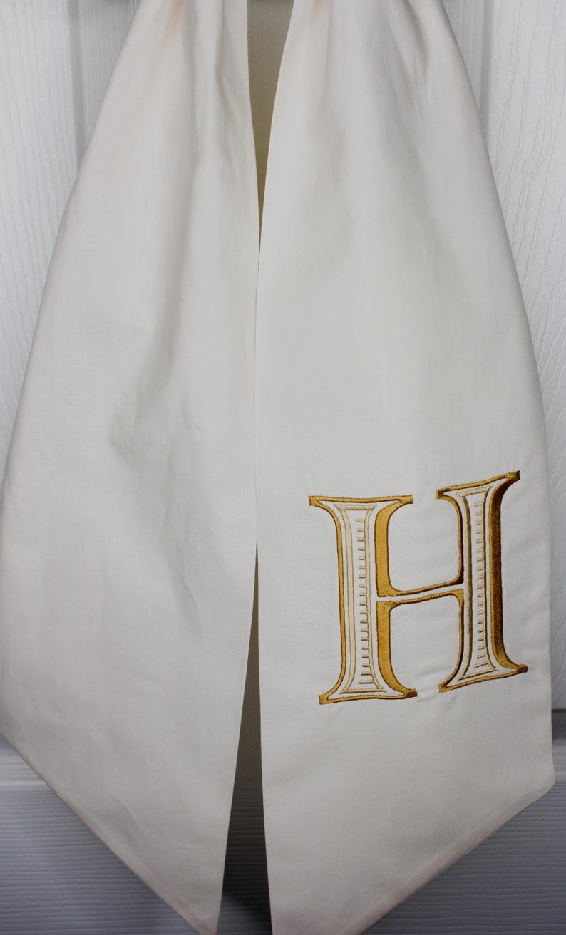 May include: A white fabric stole featuring a large, embroidered letter H. The letter is stitched in a metallic gold thread with a white interior fill and a dark outline, creating a bold, decorative monogram detail on the smooth, solid white textile.