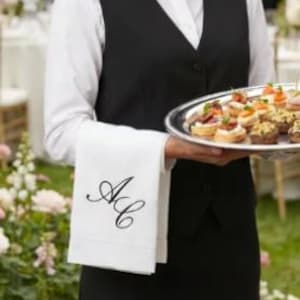 Könnte beinhalten: Eine weiße Stoffserviette mit schwarz gestickten Initialen AC, die über einem Arm liegt. Die Serviette wird neben einem silbernen Serviertablett mit verschiedenen Häppchen gehalten und mit einer schwarzen Weste und einem weißen Hemd getragen.