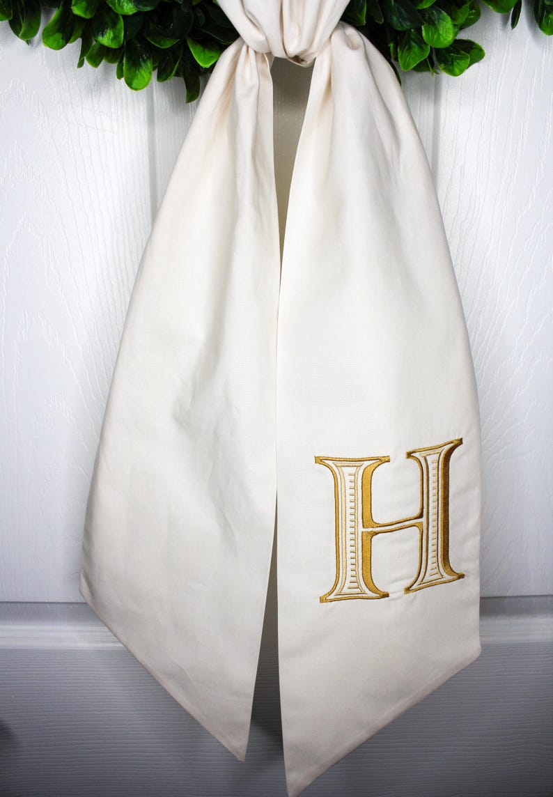 May include: An off-white fabric towel or hanging cloth featuring a large, embroidered letter H in a metallic gold thread. The letter has a serif font style with detailed stitching. The fabric is draped and gathered at the top, creating a vertical fold.