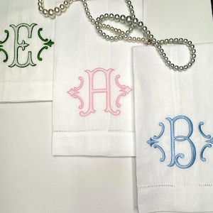 May include: Three white rectangular linen towels featuring embroidered monograms in green, pink, and blue. Each towel displays a decorative scrollwork frame around the letters E, A, and B. A string of white pearls is draped across the top of the towels.