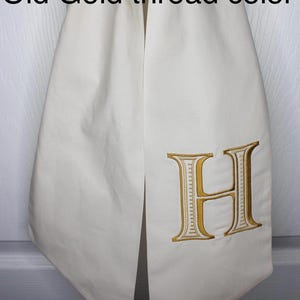 May include: A cream-colored fabric sash featuring an embroidered letter H in an old gold thread color. The text on the image reads: Cream sash, Font 1, Old Gold thread color. The sash is draped vertically with a smooth, matte finish.