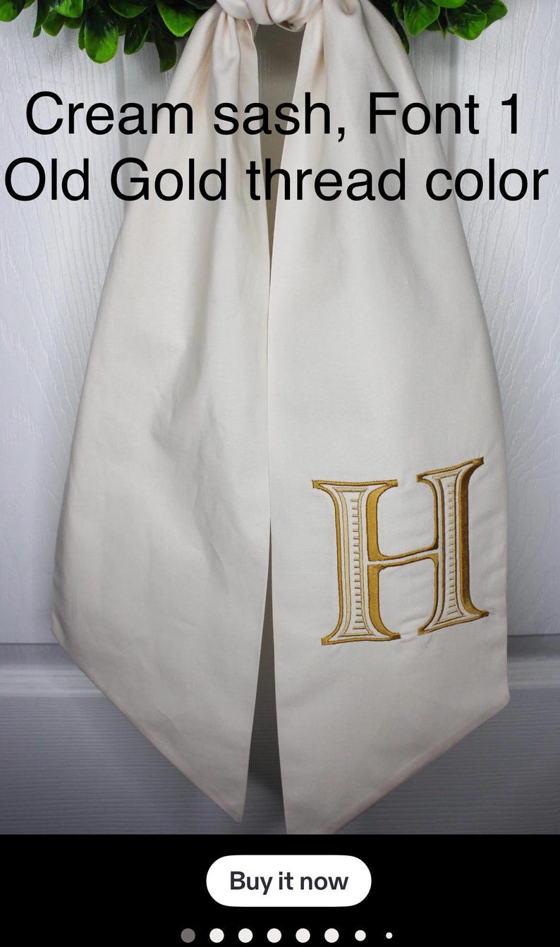 May include: A cream-colored fabric sash featuring an embroidered letter H in an old gold thread color. The text on the image reads: Cream sash, Font 1, Old Gold thread color. The sash is draped vertically with a smooth, matte finish.