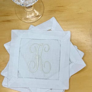 Puede incluir: Un conjunto de posavasos de lino blanco con una letra "K" bordada en el centro. Los posavasos, con un borde cosido, están apilados sobre una superficie de madera. Se ve una copa de vino de cristal.