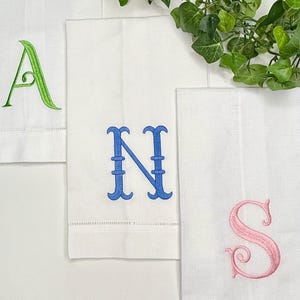 May include: Three white rectangular fabric towels featuring embroidered monograms. One towel displays a green letter A, another a blue letter N, and the third a pink letter S. Each towel has a hemstitched border detail along the bottom edge.