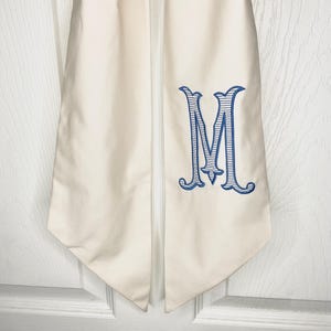 May include: A white graduation sash with a blue embroidered letter "M" in a script font.