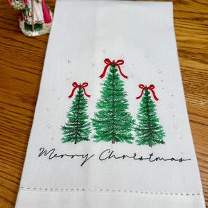 Puede incluir: Paño de cocina blanco con temática navideña con tres árboles de Navidad verdes, cada uno coronado con un lazo rojo. Las palabras "Merry Christmas" están escritas en escritura negra debajo. La toalla tiene un borde cosido decorativo.
