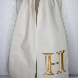 May include: A cream-colored fabric hanging decoration featuring a large, embroidered letter H in a metallic gold thread. The fabric is folded at the top and hangs in two vertical panels, displaying a clean, smooth texture and a classic serif font.