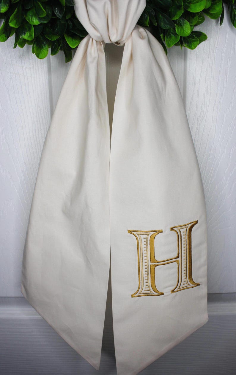 May include: A cream-colored fabric hanging decoration featuring a large, embroidered letter H in a metallic gold thread. The fabric is folded at the top and hangs in two vertical panels, displaying a clean, smooth texture and a classic serif font.