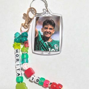 May include: A keychain featuring a rectangular clear plastic frame containing a photograph of a person in a green soccer jersey. Attached is a beaded strand with green, red, and white beads, including letter beads spelling MORITA and 19.