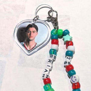 May include: A heart-shaped clear acrylic keychain featuring a photograph of a person wearing a red and white jersey. Attached is a beaded lanyard with multicolored plastic beads, including white letter beads spelling BRIAN and number beads 26.