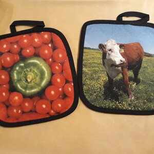 Puede incluir: Dos agarraderas de olla con dise&ntilde;os diferentes. Una agarradera de olla presenta un primer plano de tomates rojos y un pimiento verde. La otra agarradera de olla presenta una vaca marr&oacute;n y blanca de pie en un campo de hierba verde.