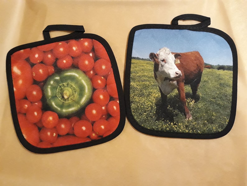 Puede incluir: Dos agarraderas de olla con dise&ntilde;os diferentes. Una agarradera de olla presenta un primer plano de tomates rojos y un pimiento verde. La otra agarradera de olla presenta una vaca marr&oacute;n y blanca de pie en un campo de hierba verde.