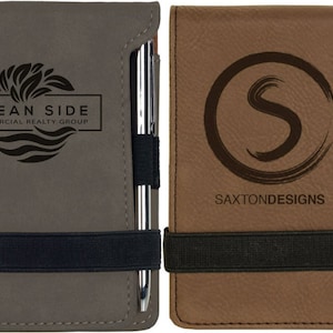 May include: Two brown leatherette journals with black elastic bands and silver pens. The left journal has the text "OCEAN SIDE COMMERCIAL REALTY GROUP" and a wave design. The right journal has the text "SAXTON DESIGNS" and a stylized letter "S" in a circle.
