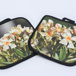 Puede incluir: Dos agarraderas de olla cuadradas en blanco y negro con un dise&ntilde;o de flores blancas. Las agarraderas tienen un borde negro.