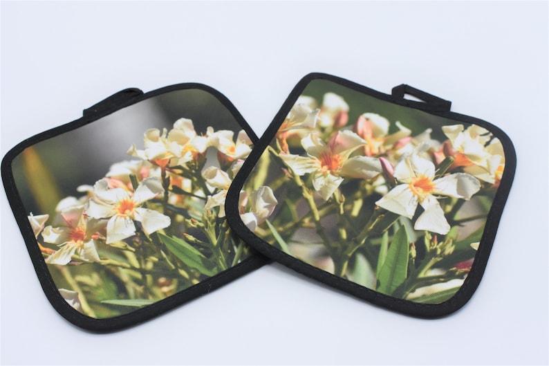 Puede incluir: Dos agarraderas de olla cuadradas en blanco y negro con un dise&ntilde;o de flores blancas. Las agarraderas tienen un borde negro.