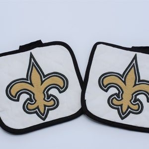 Puede incluir: Un juego de dos agarraderas de olla blancas con ribete negro y un logotipo de la flor de lis de los New Orleans Saints bordado en oro.