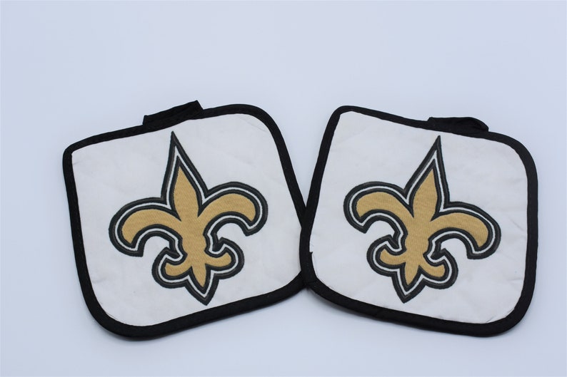 Puede incluir: Un juego de dos agarraderas de olla blancas con ribete negro y un logotipo de la flor de lis de los New Orleans Saints bordado en oro.