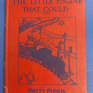 Könnte beinhalten: Ein Vintage-Exemplar von "The Little Engine That Could" von Watty Piper. Das Buchcover ist rot mit einer blauen Illustration eines Zuges und einer Strecke. Der Titel und der Name des Autors sind in weißem Text.