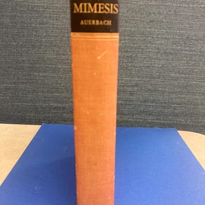 May include: A vintage book titled "Mimesis" by Auerbach. The book has a textured, orange cover with a black band at the top that displays the title and author's name in white lettering. The book is standing upright on a blue surface.