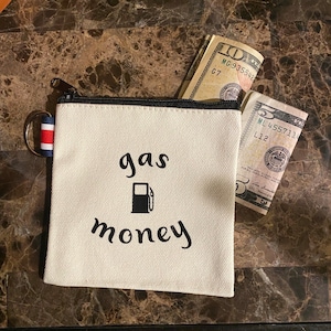 May include: A white canvas pouch with a zipper closure and a black drawstring. The pouch has the text "gas money" printed on it in black cursive font, with a black gas pump icon in between the words. The pouch is partially filled with US dollar bills.