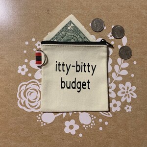 May include: A small white canvas pouch with a zipper closure. The pouch has black text that reads "itty-bitty budget". The pouch is lying on a brown surface with white floral designs.
