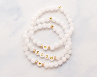 May include: Three stretch bracelets made of smooth white beads. Each bracelet features small gold-colored metallic spacer beads. One bracelet displays the word SOON in gold letters on white beads, while two others feature a single gold heart bead.