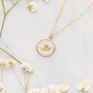 May include: A gold-toned necklace featuring a circular pendant that encases a small, pressed white flower with green stems. The pendant hangs from a delicate gold-toned chain, creating a simple and elegant piece of jewelry for any collection.