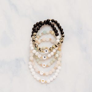 May include: Stack of beaded bracelets in various colors, including black, white, and cream. Some bracelets feature gold beads and heart-shaped charms. The bracelets are arranged on a white marble surface.