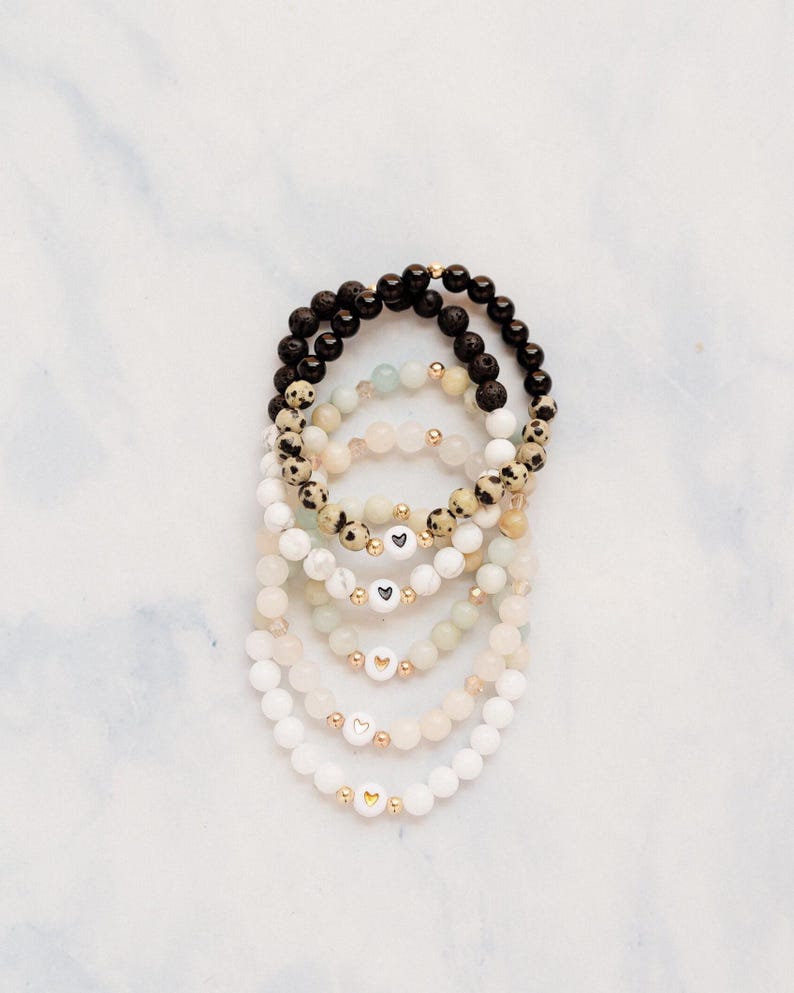 May include: Stack of beaded bracelets in various colors, including black, white, and cream. Some bracelets feature gold beads and heart-shaped charms. The bracelets are arranged on a white marble surface.