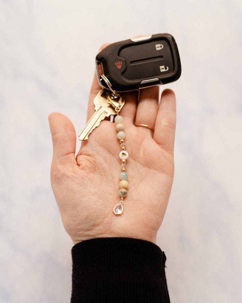 May include: A car key fob and key with a beaded keychain. The keychain features a gold-colored chain with light-colored beads, a small gold charm, and a clear teardrop-shaped charm.
