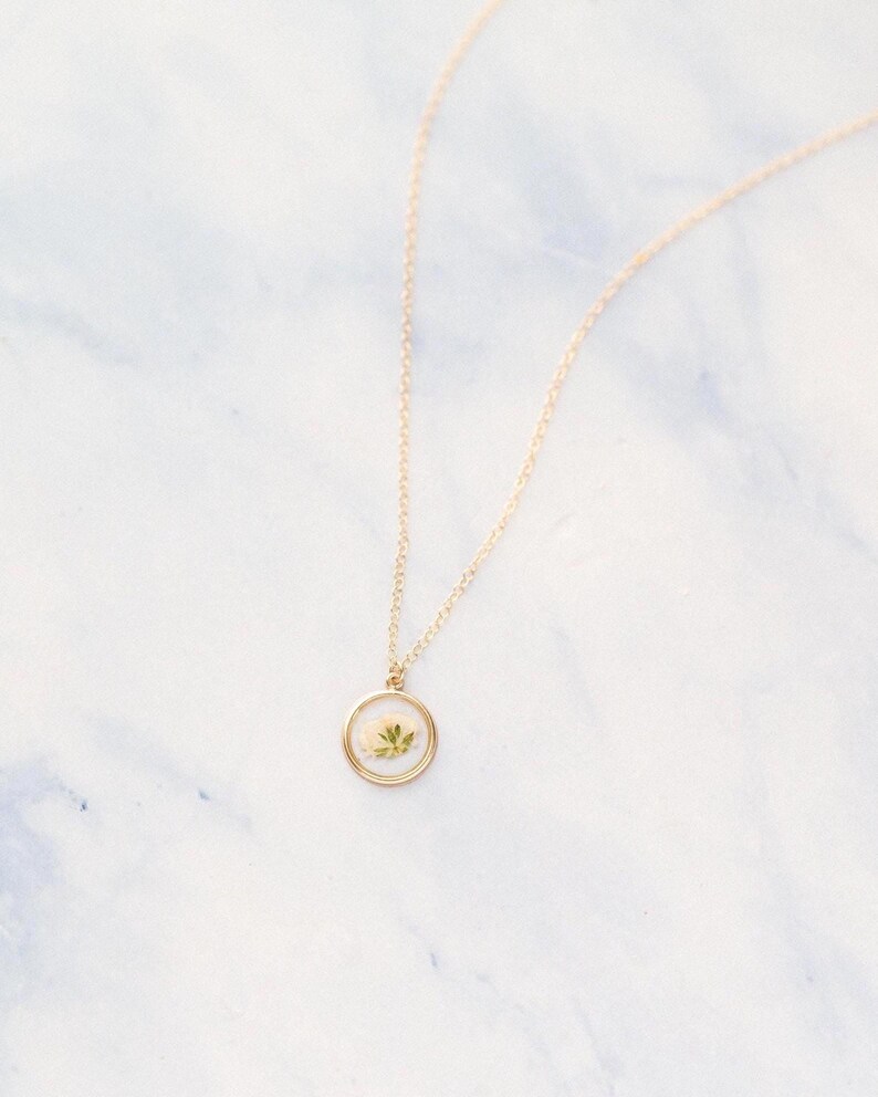 May include: A gold-toned necklace featuring a delicate chain and a circular pendant. The pendant has a thin gold rim and contains a small, pressed white flower with green leaves encased within a clear, smooth, and slightly domed resin material.