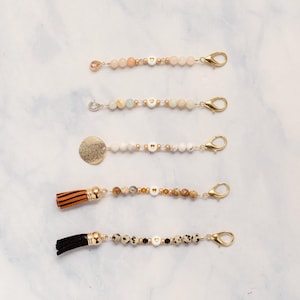 May include: Five beaded keychain charms with gold-colored clasps. The charms feature various beads, including peach, white, and speckled stones, with gold heart-shaped beads. One charm has a gold disc, and two have tassels in brown and black.