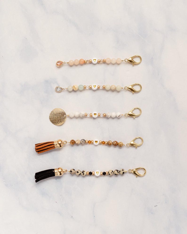May include: Five beaded keychain charms with gold-colored clasps. The charms feature various beads, including peach, white, and speckled stones, with gold heart-shaped beads. One charm has a gold disc, and two have tassels in brown and black.