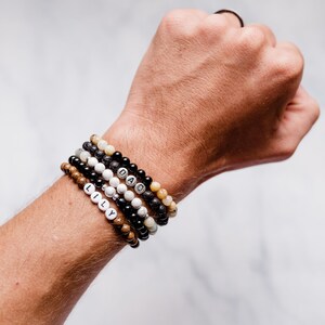 K&ouml;nnte beinhalten: Ein Stapel Perlenarmb&auml;nder mit den Worten "LILY" und "DAD" darauf. Die Armb&auml;nder bestehen aus schwarzen, braunen, wei&szlig;en und beigefarbenen Perlen.