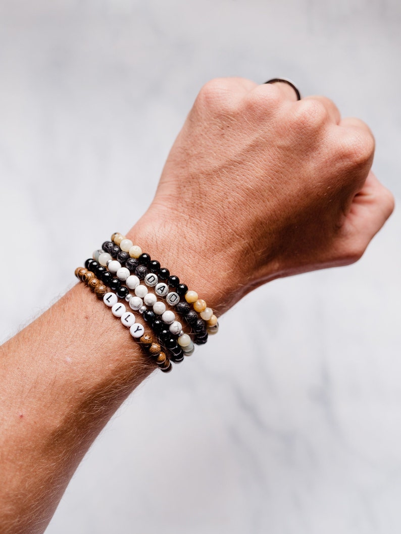 K&ouml;nnte beinhalten: Ein Stapel Perlenarmb&auml;nder mit den Worten "LILY" und "DAD" darauf. Die Armb&auml;nder bestehen aus schwarzen, braunen, wei&szlig;en und beigefarbenen Perlen.