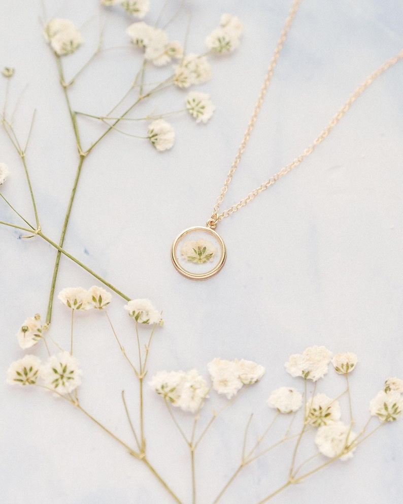 May include: A gold-toned necklace featuring a circular pendant that encases a small, pressed white flower with green stems. The pendant hangs from a delicate gold-toned chain, creating a simple and elegant piece of jewelry for any collection.