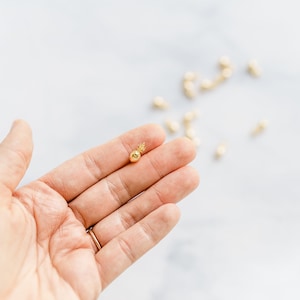 Puede incluir: Un pequeño dije de piña de color dorado se sostiene en la mano de una persona. El dije tiene una superficie texturizada y una corona detallada. Otros dijes similares están dispersos en el fondo sobre una superficie de mármol blanco.