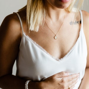 May include: A gold chain necklace with a small circular pendant containing a pressed flower, a double-strand white beaded bracelet with gold accents, and a thin gold ring. The jewelry is worn with a white camisole featuring a frayed lace trim.