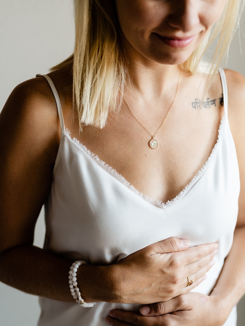 May include: A gold chain necklace with a small circular pendant containing a pressed flower, a double-strand white beaded bracelet with gold accents, and a thin gold ring. The jewelry is worn with a white camisole featuring a frayed lace trim.