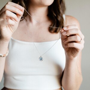 May include: A silver necklace with a small, round pendant containing a blue and white design. The necklace is being held up by a person wearing a white tank top. A bracelet with light-colored beads is visible on the left wrist.