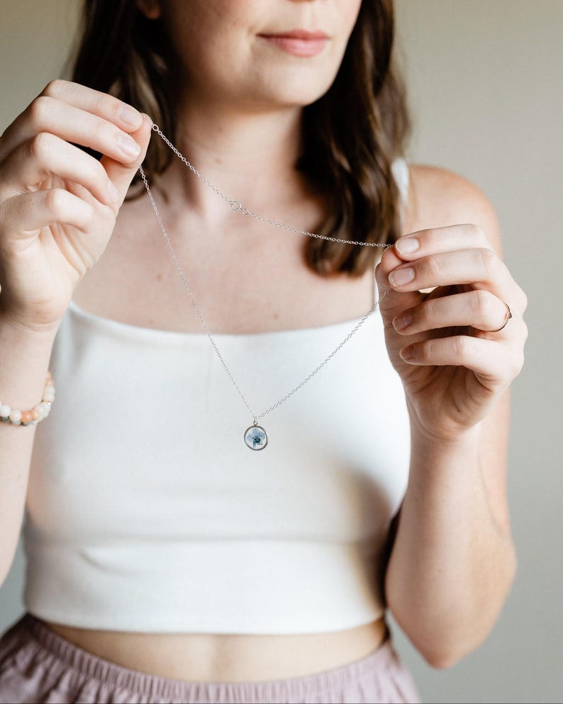 May include: A silver necklace with a small, round pendant containing a blue and white design. The necklace is being held up by a person wearing a white tank top. A bracelet with light-colored beads is visible on the left wrist.