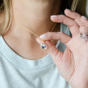 May include: A gold-colored necklace with a delicate chain and a round pendant. The pendant features a small, encased floral design with blue and white hues. The necklace is being held up, showcasing the pendant's details.