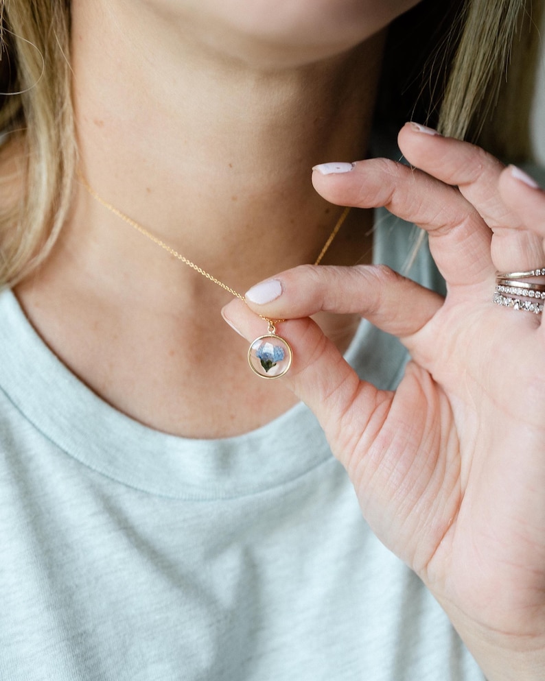 May include: A gold-colored necklace with a delicate chain and a round pendant. The pendant features a small, encased floral design with blue and white hues. The necklace is being held up, showcasing the pendant's details.