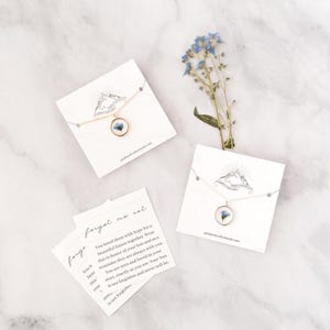 May include: A gold-tone necklace with a round pendant containing a blue flower, displayed on a white card. The card has a mountain logo and the text "peakandvalleybeads.com". A sprig of blue flowers and a card with text "forget me not" are also present.