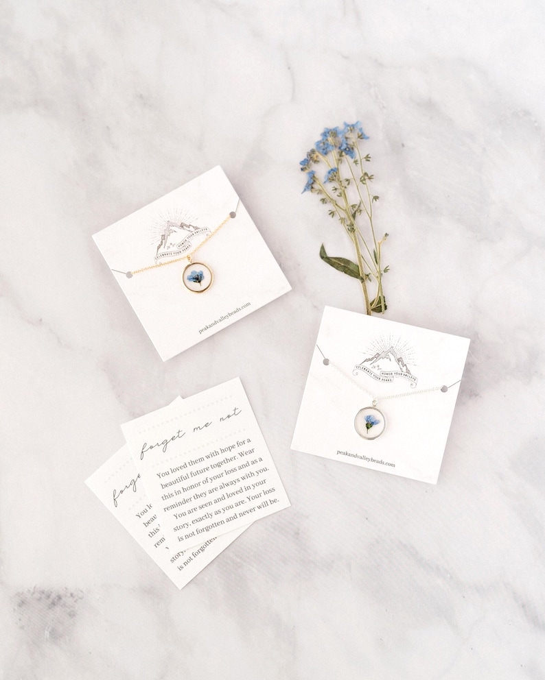 May include: A gold-tone necklace with a round pendant containing a blue flower, displayed on a white card. The card has a mountain logo and the text "peakandvalleybeads.com". A sprig of blue flowers and a card with text "forget me not" are also present.
