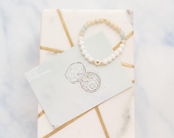 May include: A beaded bracelet featuring white and pale blue spherical beads, a small gold-toned bead, and a central white bead with a gold heart symbol. The bracelet rests on a rectangular white marble box with gold geometric lines and a card.