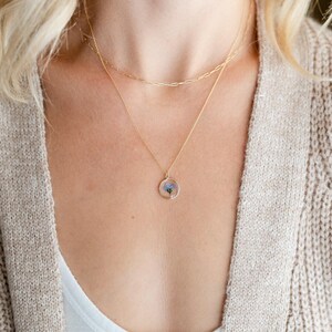 May include: A gold-colored necklace with three layers. The bottom layer features a circular pendant with a small blue flower encased in clear resin. The other two layers are delicate chains. The person is wearing a white top and a beige cardigan.
