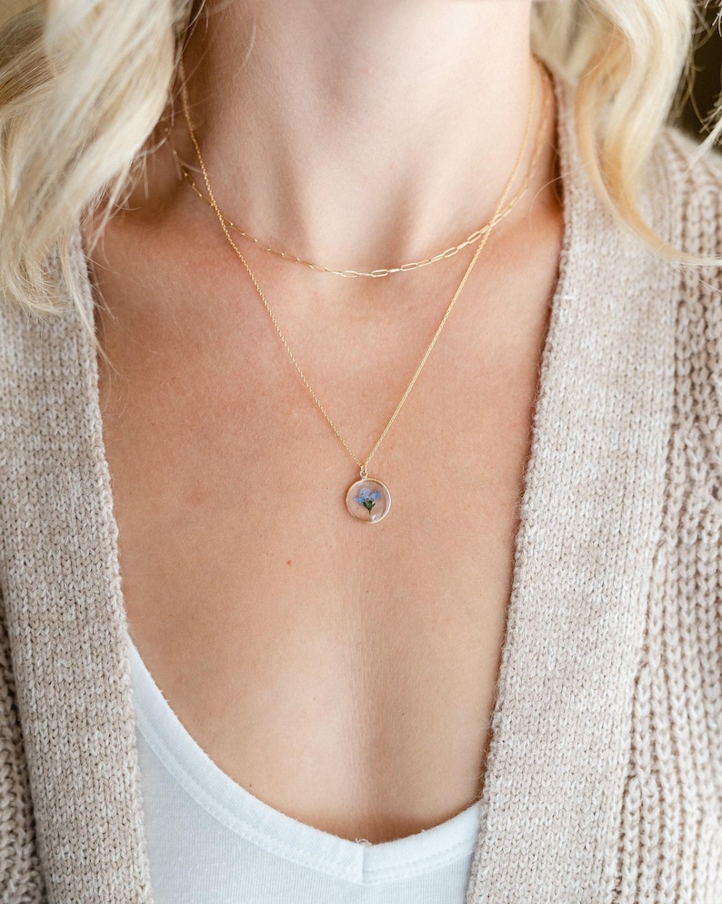 May include: A gold-colored necklace with three layers. The bottom layer features a circular pendant with a small blue flower encased in clear resin. The other two layers are delicate chains. The person is wearing a white top and a beige cardigan.